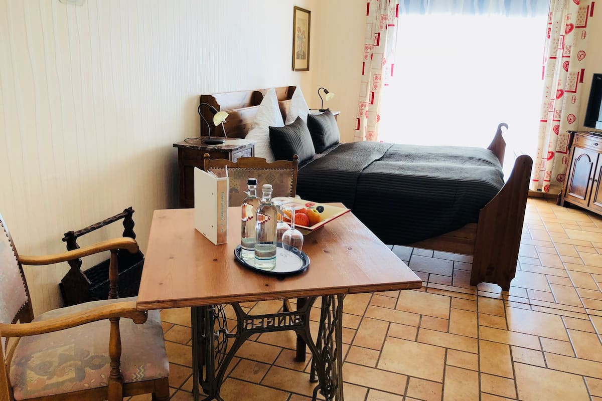 Una habitación acogedora con una mesa de madera, una máquina de coser, una cama con una manta oscura y una ventana con cortinas.