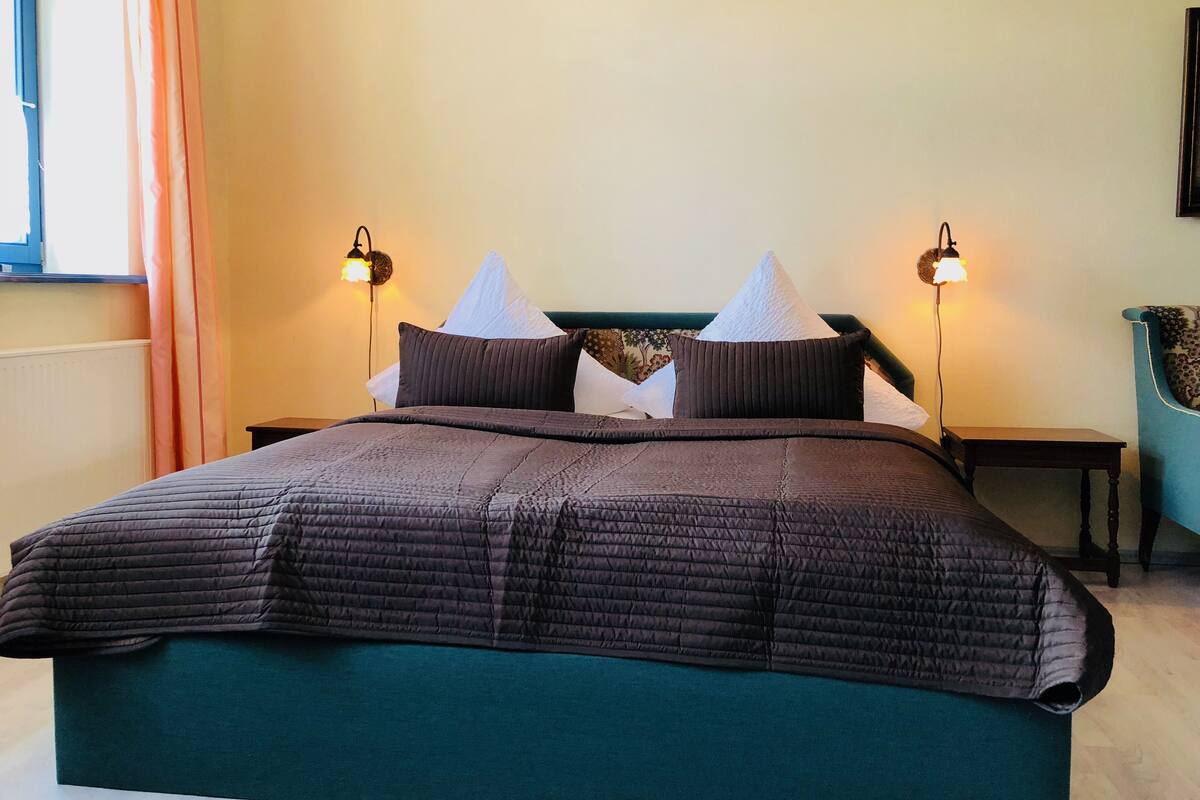 Una cama bien hecha con un edredón oscuro, dos lámparas de noche y una ventana con cortinas.
