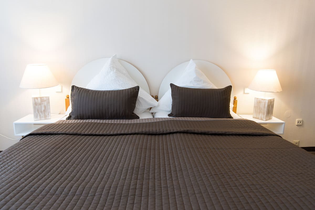 Una cama bien hecha con un edredón marrón, dos almohadas y dos lámparas de noche sobre mesitas blancas.