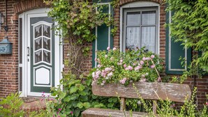 Een bakstenen huis met een groene deur en luiken, een houten bankje en roze bloemen.