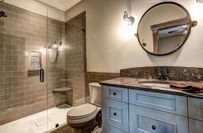 Et badeværelse med en glasafskærmet brusekabine, toilet, vask med skiferplade og et spejl med lysarmatur.