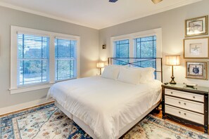 A bedroom with a large bed, two bedside tables, and a nightstand. There are two windows with blinds, a wall-mounted cross, and framed pictures. - Corpus Christi Home w/ Pool & Hot Tub: Walk to Bay (Corpus Christi)