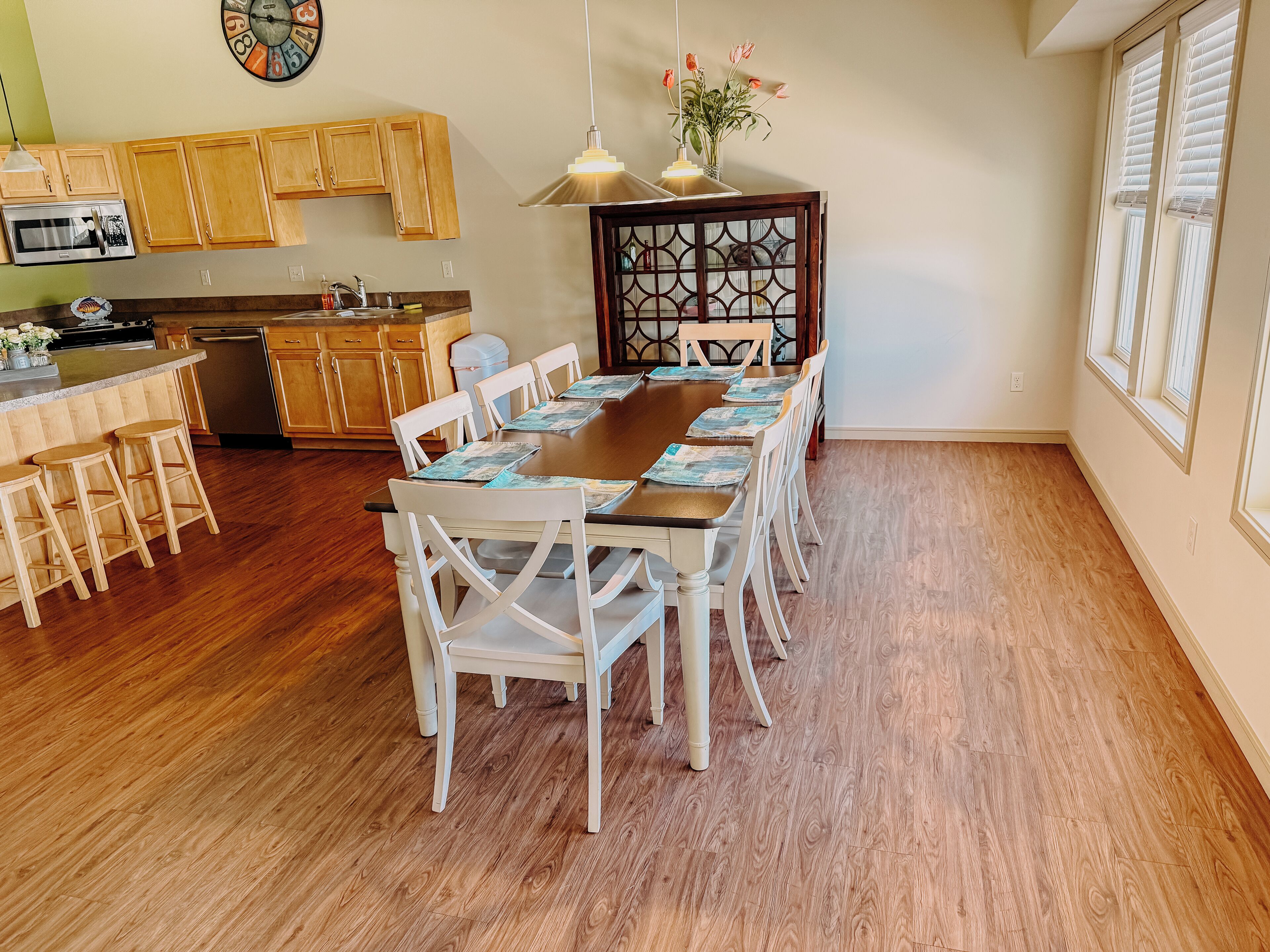 Un comedor con una mesa de madera y sillas blancas, una cocina con armarios de madera y un reloj en la pared.