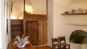 A wooden dining table with a floral centerpiece, two wooden chairs, a white curtain, and a shelf with decorative plates.