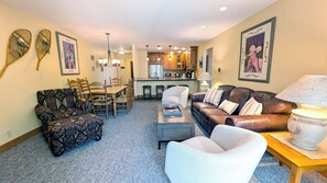 A living room with a brown leather sofa, patterned armchair, and a white armchair. There is a dining table with chairs, a kitchen in the background, and artwork on the walls.