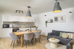 Une cuisine moderne avec des armoires blanches, un four à micro-ondes et un grille-pain. On y trouve une table à manger avec des chaises et un vase décoré de branches. Dans le salon, il y a un canapé gris et une petite table ronde sur laquelle sont posés un livre et un bol.
