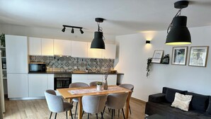 A modern kitchen with white cabinets, a microwave, and a patterned backsplash. There is a dining table with chairs and a black pendant light above it. A black sofa and a small table are in the living area.