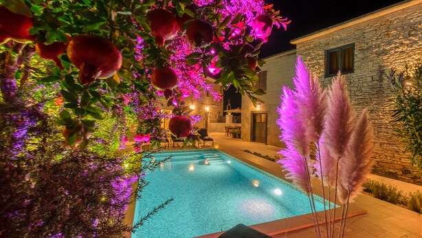 A swimming pool with a stone wall and outdoor seating area, surrounded by potted plants and decorative lighting.