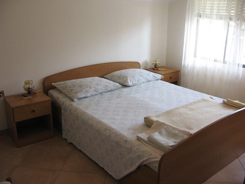 Uma cama bem arrumada com colcha estampada e almofadas, cabeceira de madeira, mesa de cabeceira com abajur e janela com cortinas leves.