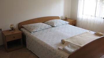 Uma cama bem arrumada com colcha estampada e almofadas, cabeceira de madeira, mesa de cabeceira com abajur e janela com cortinas leves.