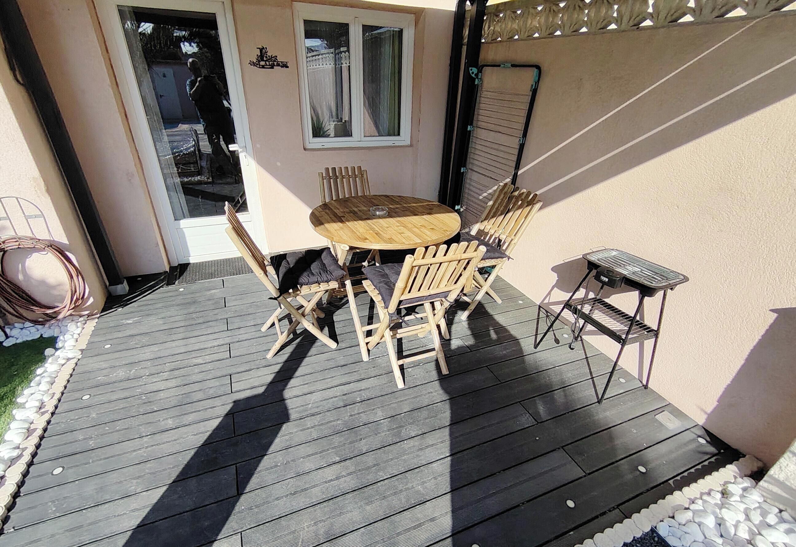 Eine Holzterrasse mit einem runden Tisch, Stühlen, einem Grill und einer Schiebetür.
