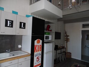 Eine kompakte Küche mit weißen Schränken, einer Spüle, einer Mikrowelle und einem Kühlschrank. Auf den Schränken befinden sich Schilder und ein Werbeschild für 'KUB' am Kühlschrank.