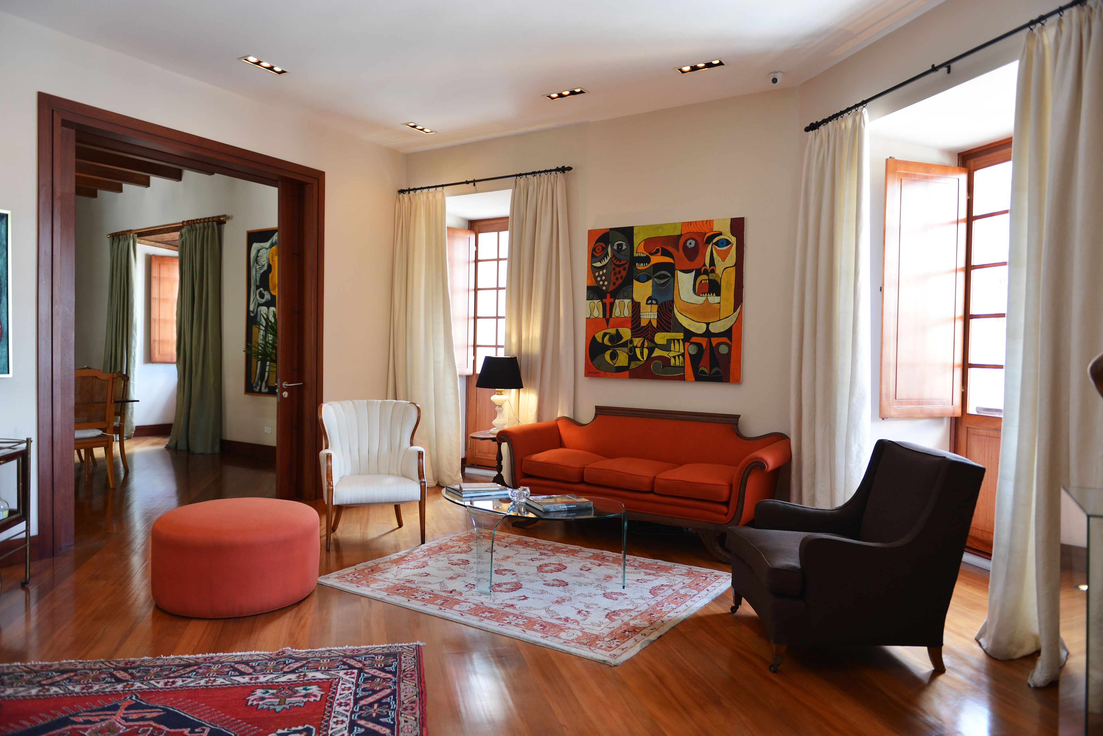 Amplia sala de estar con piso de madera, un sofá rojo, un sillón negro y un sillón blanco. Hay un cuadro grande en la pared y una ventana con cortinas.