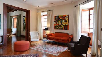 Amplia sala de estar con piso de madera, un sofá rojo, un sillón negro y un sillón blanco. Hay un cuadro grande en la pared y una ventana con cortinas.