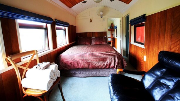 A cozy bedroom with a bed, a wooden chair with towels, a leather couch, and a window with curtains. - Waitomo Express - 1918 Train Carriage (Waitomo)