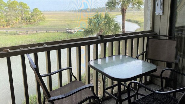 A balcony with a table and chairs, overlooking a river and fields. - 167 Beach Club Villa - Marsh & Canal View - Golf Cart Included (Fripp Island)