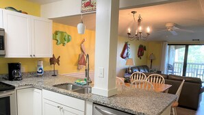 A kitchen with white cabinets, granite countertops, and a dishwasher. There is a dining area with a table and chairs, and a view of the outdoors through a large window. - 167 Beach Club Villa - Marsh & Canal View - Golf Cart Included (Fripp Island)