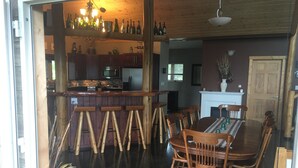 A dining area with a wooden table and chairs, a bar with stools, and a kitchen in the background. - Stunning Post and Beam Home with the best views in the valley, wheelchair acc. (Penticton)