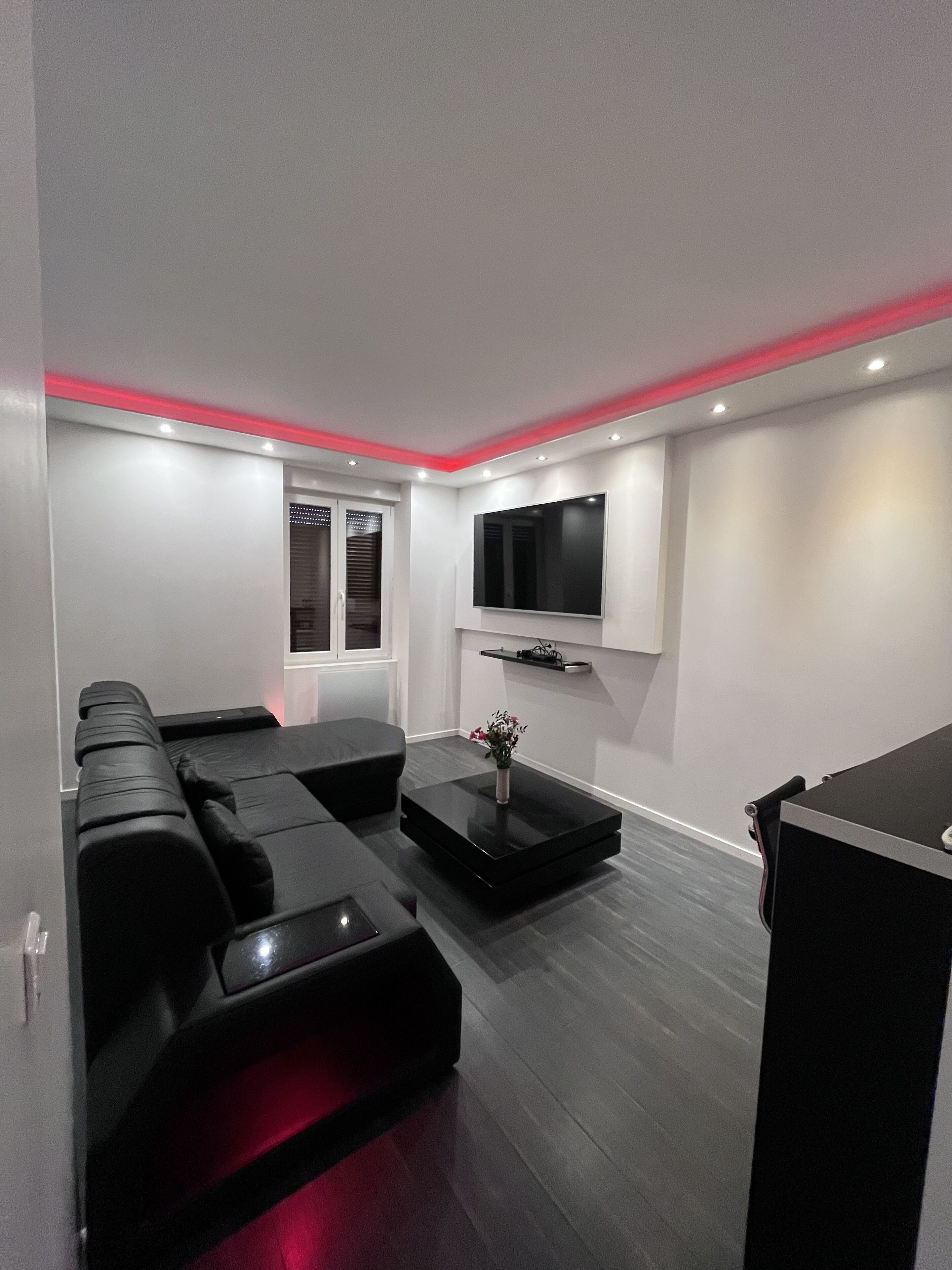 Een moderne woonkamer met een zwarte hoekbank, een bijzettafeltje met een vaas en een flatscreen televisie aan de muur.