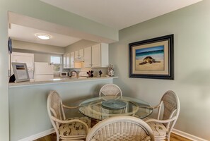 A dining area with wicker chairs and a glass table, a kitchen with white cabinets, and a framed picture of a beach scene on the wall.