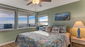 A bedroom with a large bed, a ceiling fan, a nightstand, and a view of the ocean through the windows.