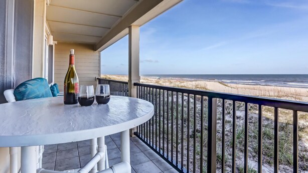 A round table with a bottle of wine, two glasses, and a candle on a balcony overlooking a beach.