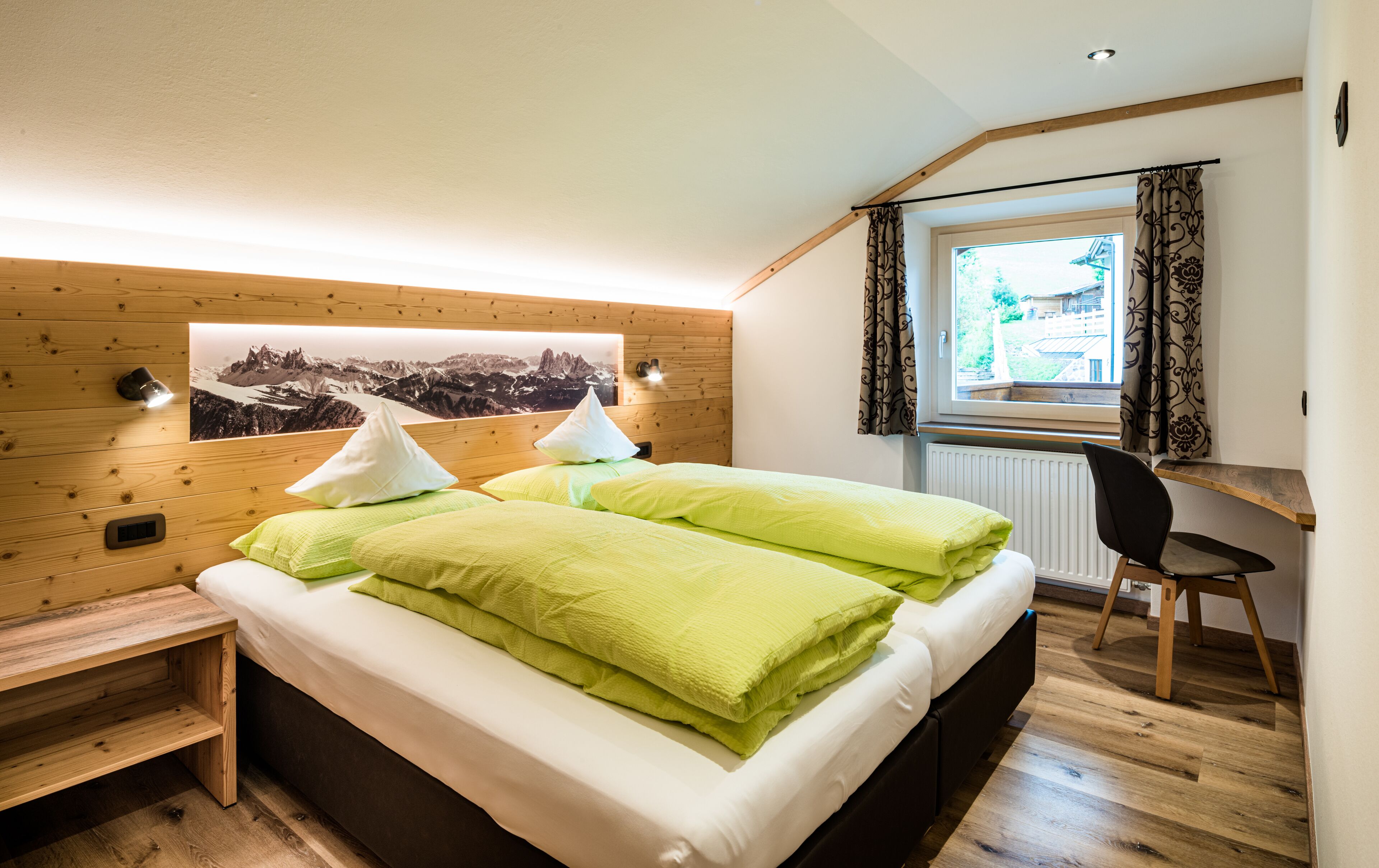 Une chambre avec une tête de lit en bois, un lit avec des oreillers verts, une table de chevet, un bureau avec une chaise, une fenêtre avec des rideaux et un tableau accroché au mur.