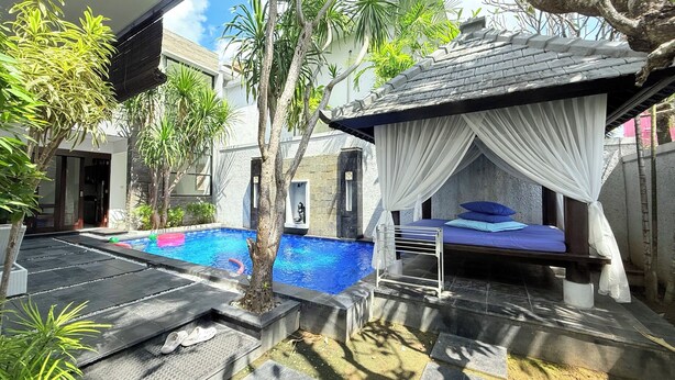 Un área de piscina moderna al aire libre con una cama bajo una pérgola, rodeada de plantas tropicales y un sendero de piedra.