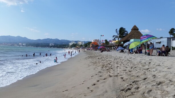 Uma praia com pessoas nadando e caminhando na orla, guarda-sóis e edifícios ao fundo.