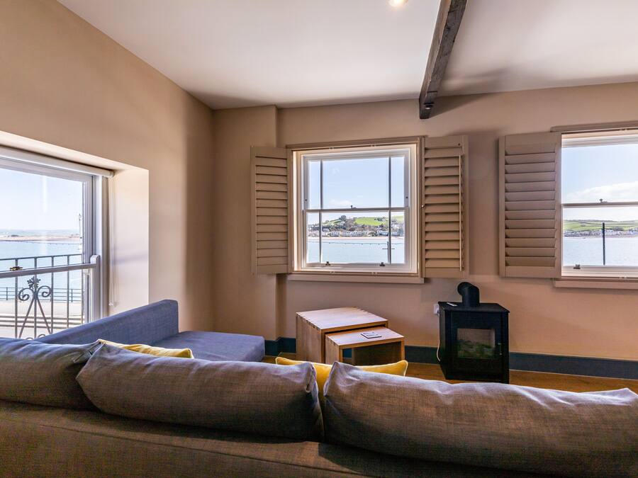A room with a sofa, a small table, and a heater. There are three windows with shutters, and a balcony with a railing.