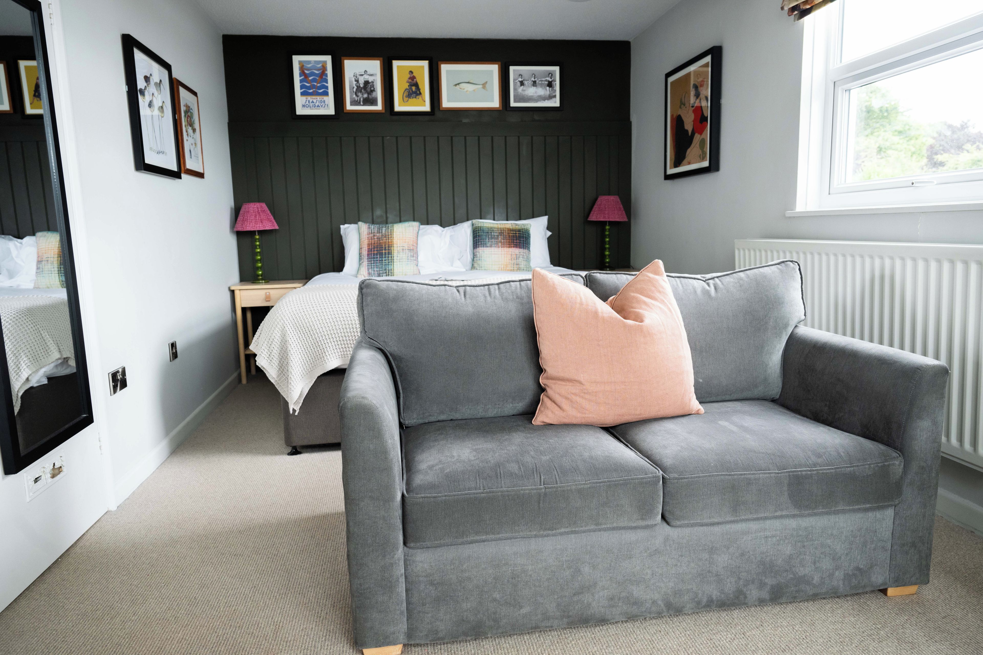 Un dormitorio moderno con un sofá gris, una cama con muchas almohadas, una mesita de noche con lámparas y cuadros enmarcados en la pared.