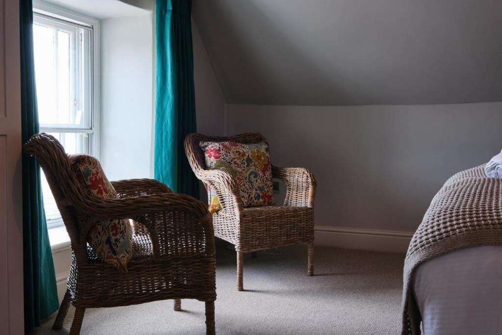 A room with two wicker chairs, one with a floral cushion and another with a patterned cushion, a window with curtains, and a carpeted floor.