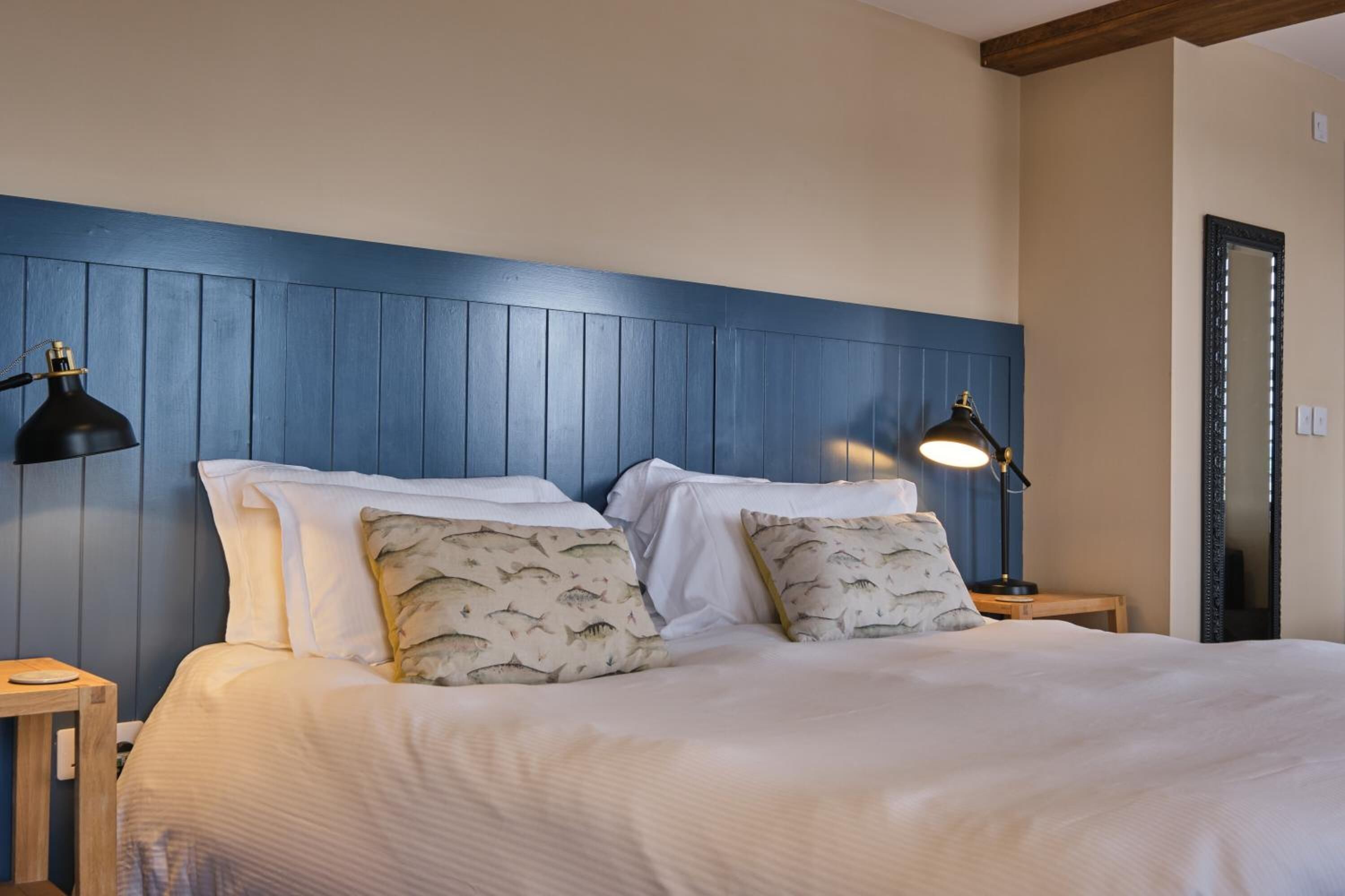 A neatly made bed with white linens and patterned pillows, flanked by two bedside tables with lamps, in a room with blue paneled walls and a wooden ceiling.