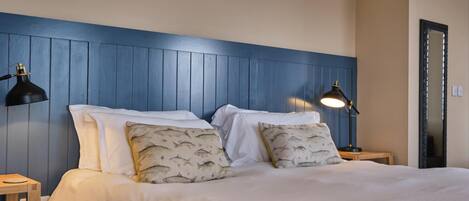 A neatly made bed with white linens and patterned pillows, flanked by two bedside tables with lamps, in a room with blue paneled walls and a wooden ceiling.