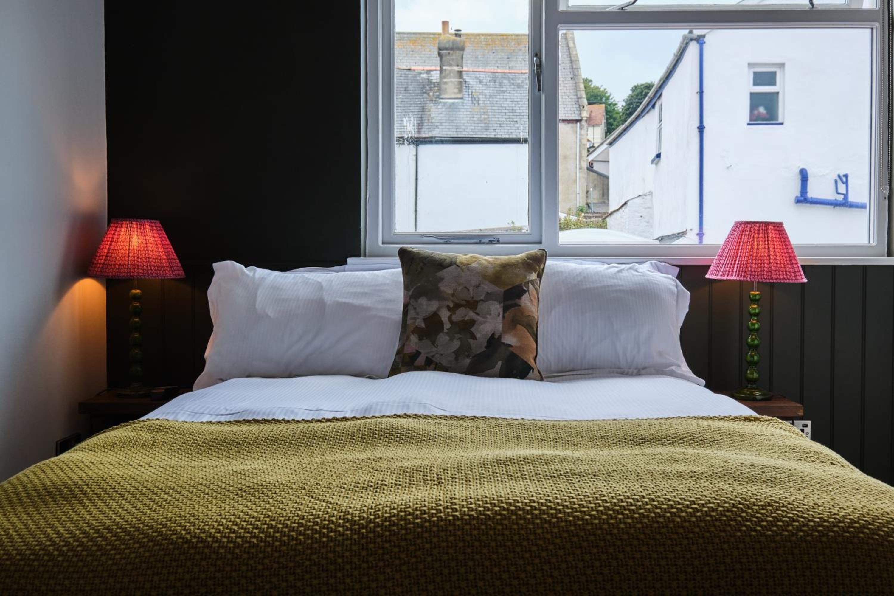A bed with white linens and a green knitted blanket, flanked by two lamps with red shades, in a room with a window overlooking a white building.