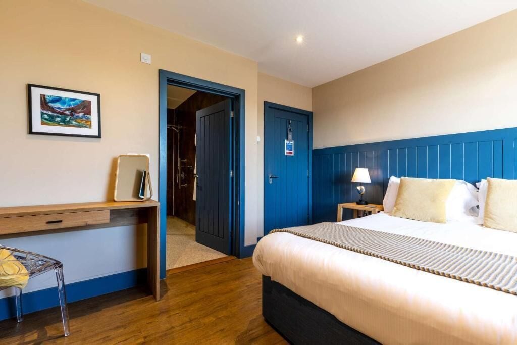 A hotel room with a bed, a desk, a chair, a nightstand, and a door.