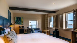 Een slaapkamer met een groot bed, een bureau, een stoel, een schilderij en uitzicht op zee door de ramen.