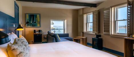 A bedroom with a large bed, a desk, a chair, a painting, and a view of the sea through the windows.
