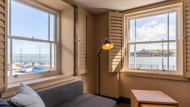Een kamer met een grijze bank, een houten tafel en twee ramen met uitzicht op een waterpartij en een brug.