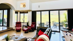 A spacious living room with a pool table, red leather chairs, and large windows offering a view of the outdoors.