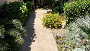 A pathway flanked by lush greenery and flowering plants leading to a building with a tiled roof and a large window.