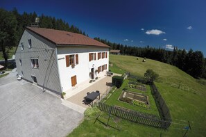 Une maison à deux étages, avec un toit recouvert de tuiles rouges, un jardin clôturé et une vue dégagée sur la forêt environnante.