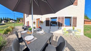 An outdoor dining area with a table set for four, surrounded by wicker chairs, under a large umbrella. - Marie-Lou's Farm - La Joux Dessus (Prémanon)