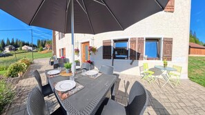 Un espace de restauration en plein air avec une table dressée pour quatre personnes, entourée de chaises en osier, sous un grand parasol.