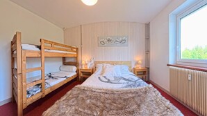 Een kamer met stapelbedden, een groot bed met een deken, een bont tapijt, een muur met een bloemig kunstwerk, een raam en een radiator.
