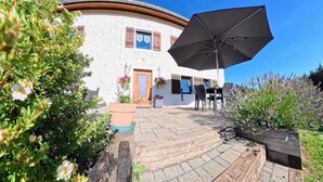 A white building with a curved roof, a wooden door, and a large black umbrella. There are potted plants and a table with chairs outside. - Marie-Lou's Farm - La Joux Dessus (Prémanon)