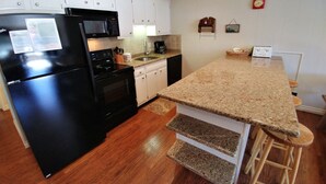 Une cuisine de taille réduite avec des meubles blancs, un réfrigérateur noir, un four à micro-ondes, une cuisinière et un évier. On y trouve un tabouret en bois et un petit tapis sur le sol.