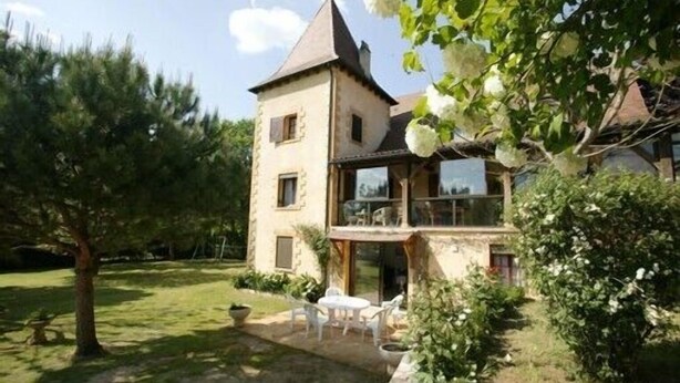 A two-story stone house with a balcony, surrounded by a well-maintained lawn and trees.