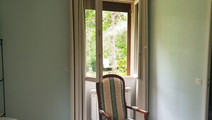 A room with a wooden chair with a patterned cushion, a table with a white tablecloth, and a window with curtains.