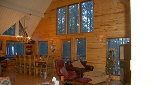 A spacious cabin with a high ceiling, large windows, a dining area, a living room with a fireplace, and a Christmas tree.